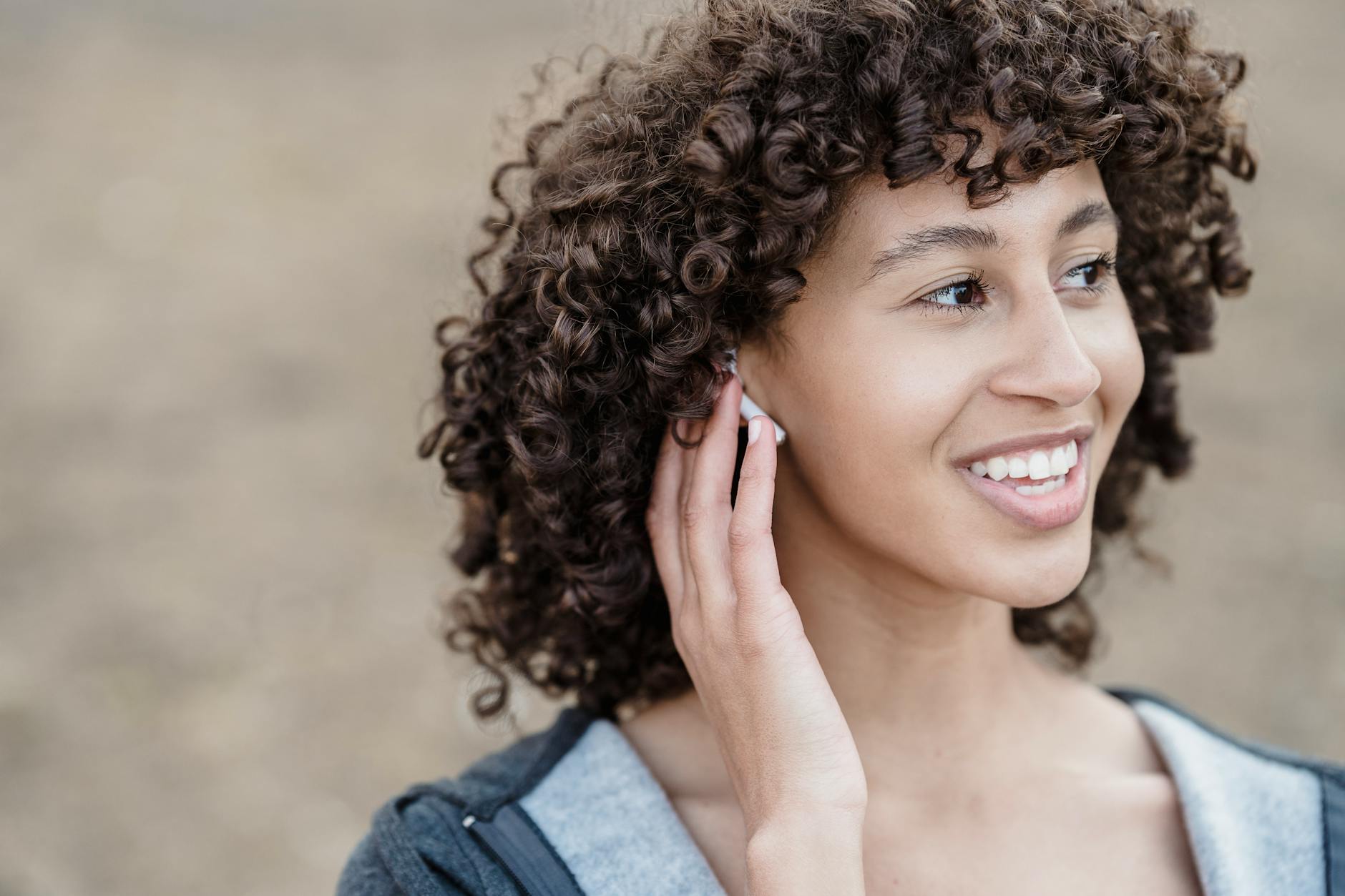 Cheerful ethnic female with black curly hair looking away with smile while listening to songs in wireless earphones on street against blurred background